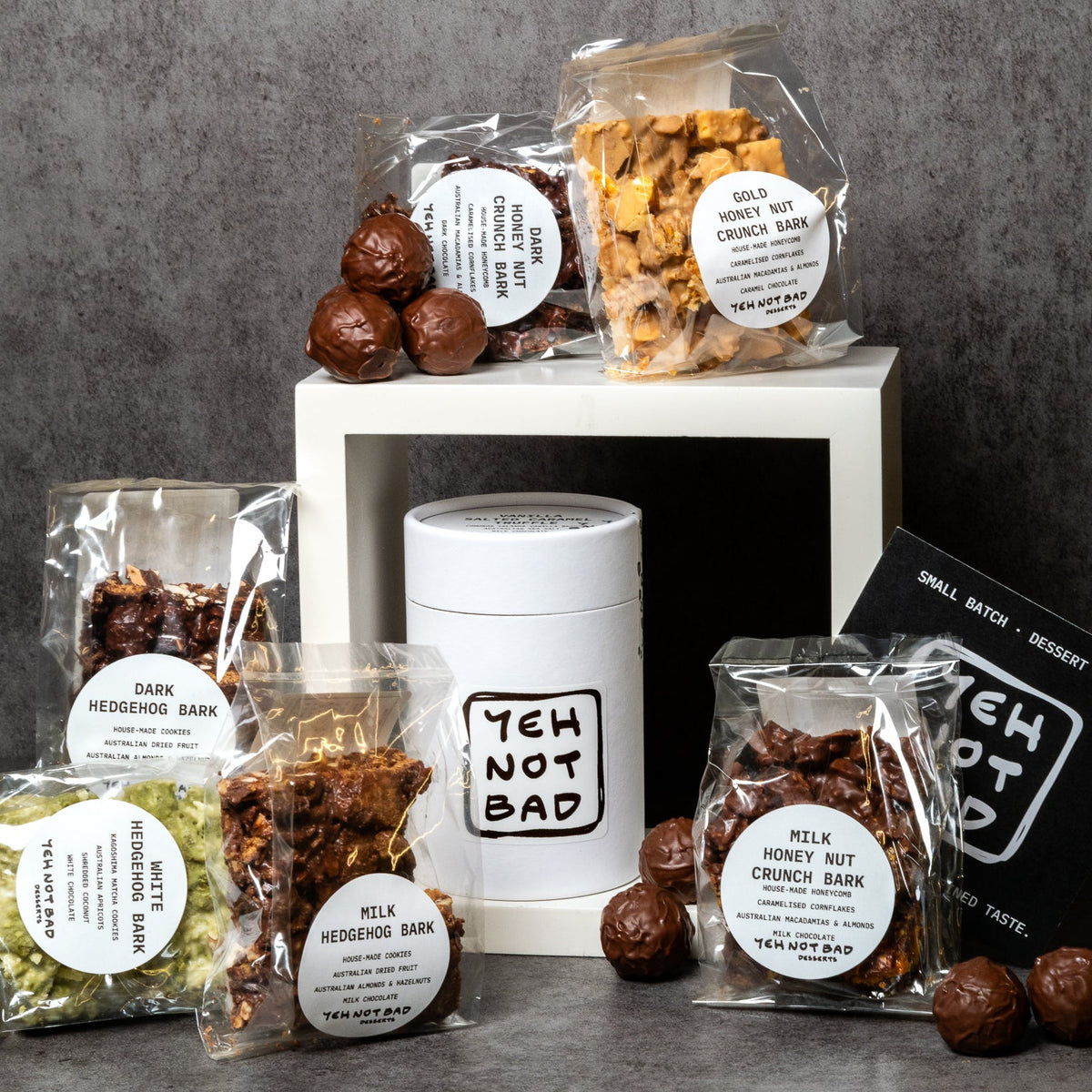 A curated assortment of tea and snack products from Yeh Not Bad, including various packaged dark and milk herbal/fruit bars, honey nut crisp bars, and a white canister, arranged on a white pedestal against a textured gray background.