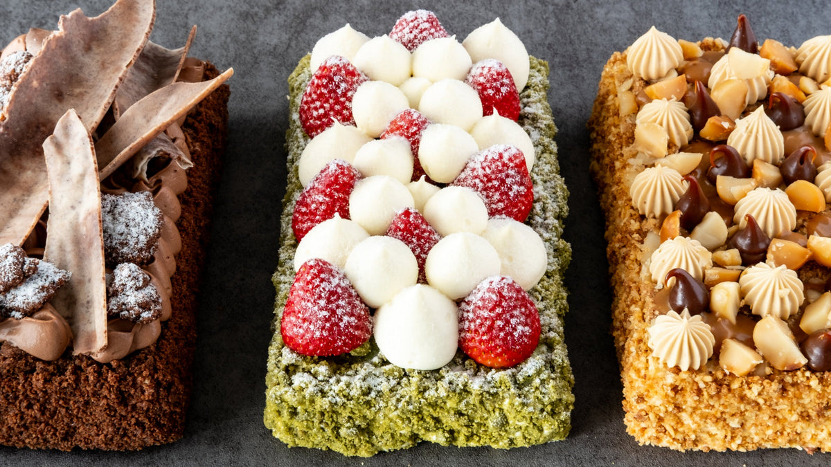 Three decorated rectangular cakes arranged side by side on a dark gray surface. The left cake is chocolate with brown chocolate shavings and powdered sugar.