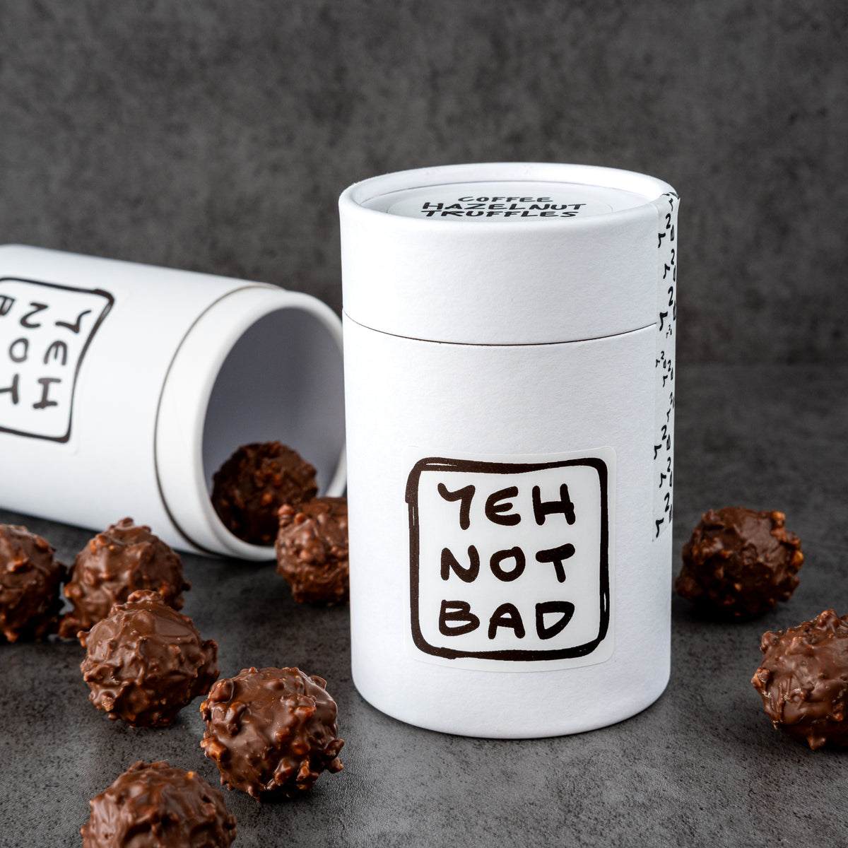 A white cylindrical chocolate tin with the phrase 'YEH NOT BAD' printed in black on the front, standing upright on a dark gray surface with several chocolate-covered candies scattered around it. A second tin lies on its side in the background.