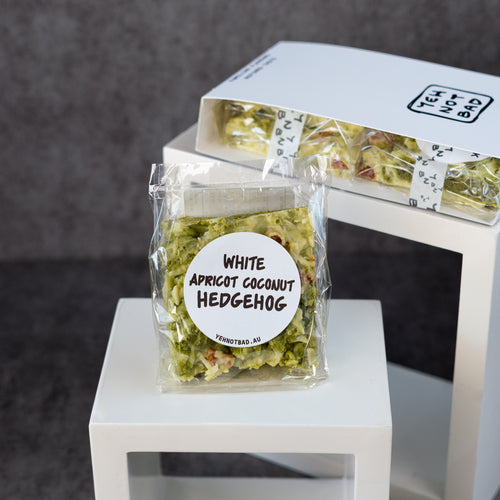 A packaged food product labeled 'WHITE APRICOT COCONUT HEDGEHOG' by Yeh Not Bad, shown in a clear bag with a round white label, alongside a white branded box on white display risers against a dark gray background.