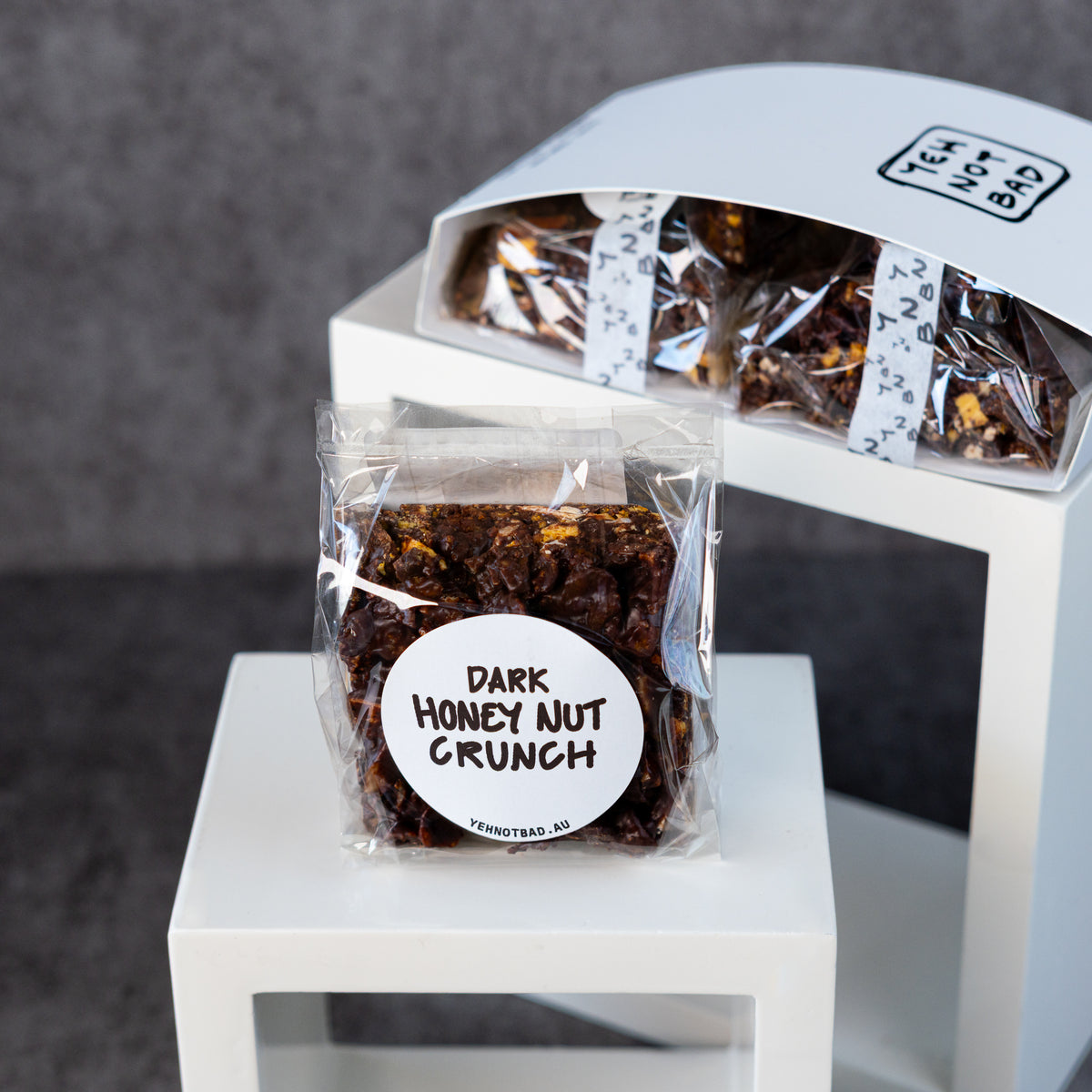 A clear bag of dark honey nut crunch with a white circular label sits on a white pedestal, with a matching box of the same snack behind it on a gray background.