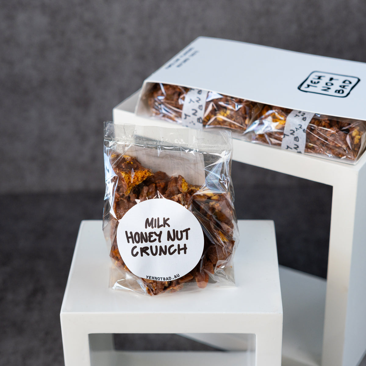 A clear plastic bag of milk honey nut crunch sits on a white pedestal, with a matching white box of the same product displayed behind it against a dark gray background.