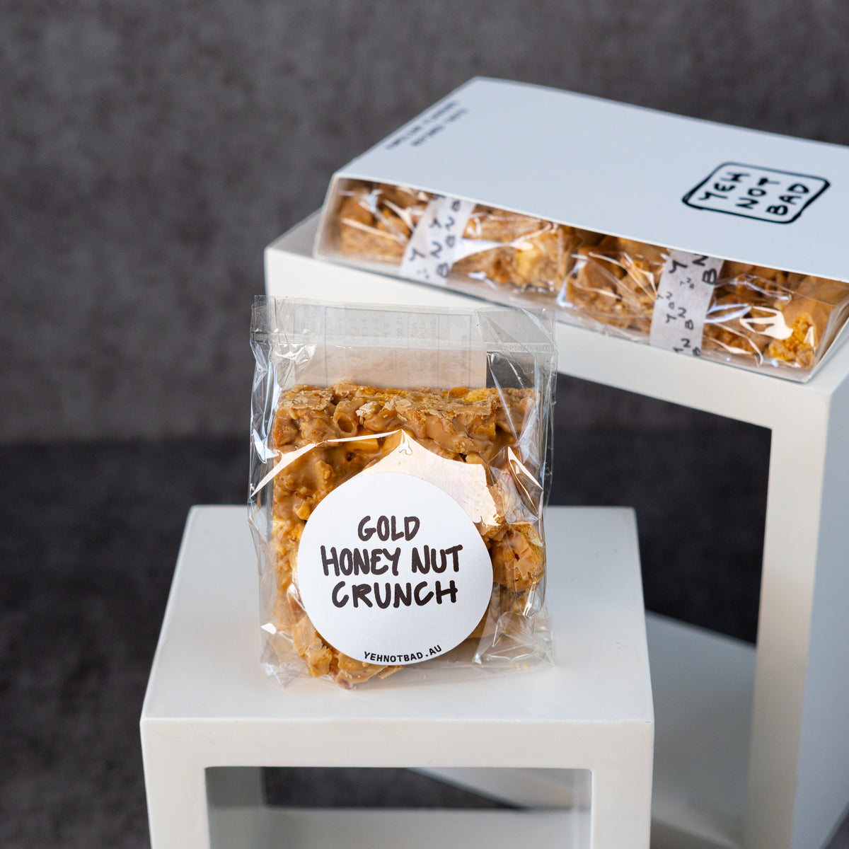 A clear bag of Gold Honey Nut Crunch cereal clusters sits on a white pedestal in front of a matching white box of the same product, photographed against a dark gray textured background.