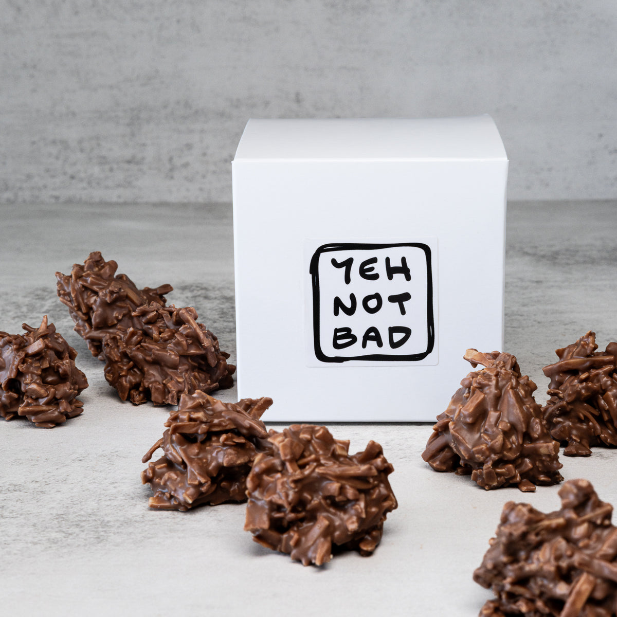 A white square box with a black hand-drawn style logo reading 'YEH NOT BAD' sits on a light gray surface, surrounded by several chocolate clusters. The product appears to be the boxed item or packaged food product, photographed in a clean studio setup with no people or real environment visible.