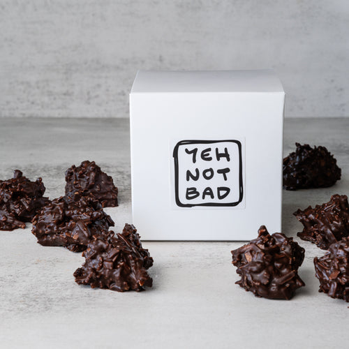 A white square box with a black sticker reading 'YEH NOT BAD' sits on a light gray surface, surrounded by several chocolate clusters.